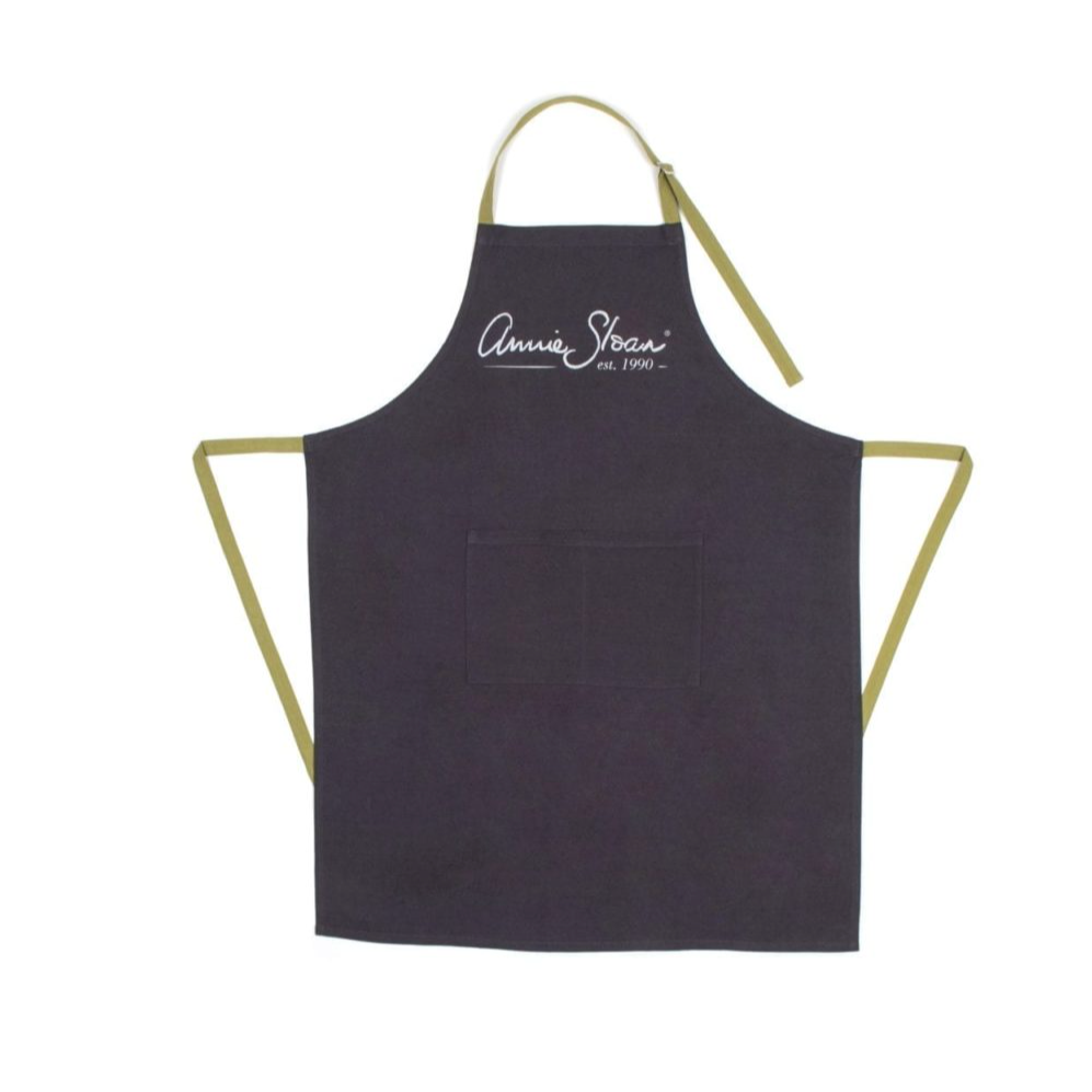 A product listing page for an Annie Sloan apron, showing a dark navy apron with gold trim on the right side, alongside product details and a checkout status message. The image is captured from a web browser.