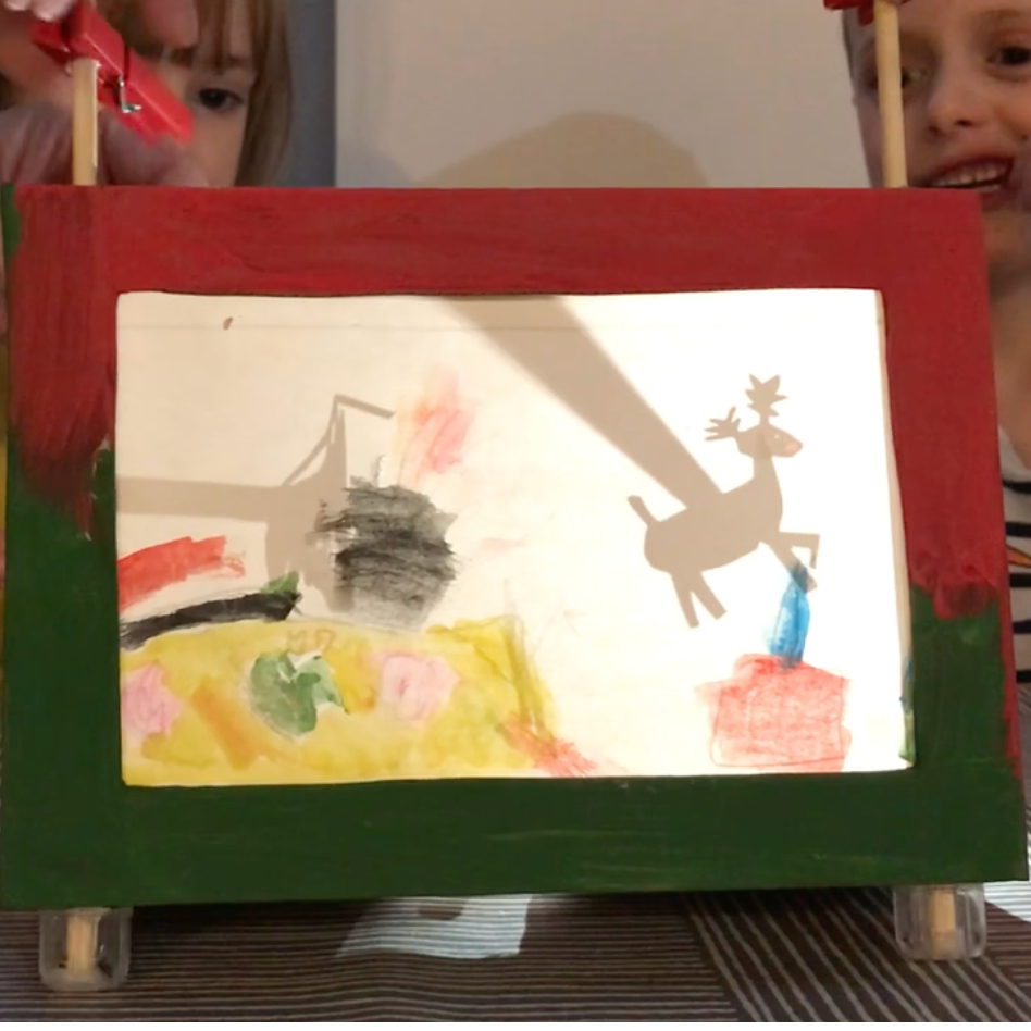 A child's painted wooden picture frame displayed on a small easel stand, with two children partially visible behind it in a home setting.
