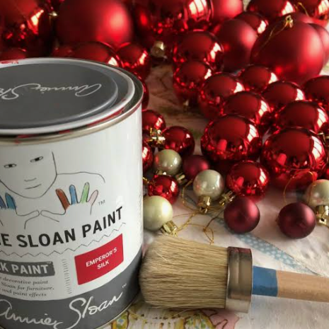 A can of Annie Sloan Paint in the color 'Emerald Red' is shown alongside a paintbrush and a background of red Christmas ornaments. Text below reads 'How to: Annie Sloan colour block.