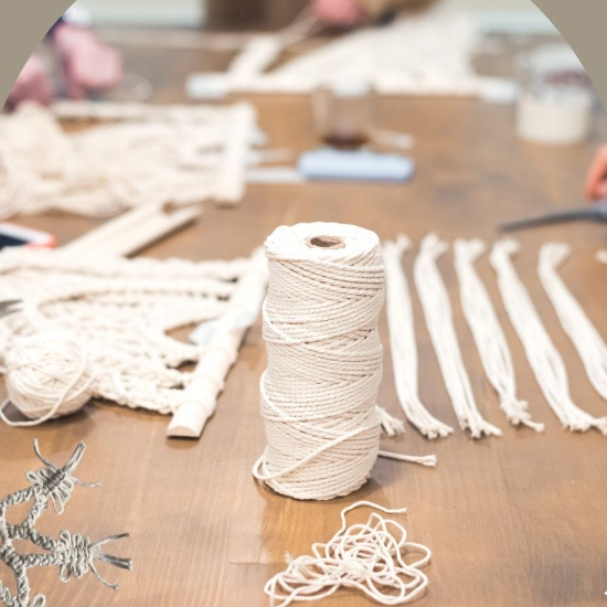 A spool of off-white cotton cord for macrame or wall hanging sits on a wooden table, surrounded by loose strands of the same cord, a partially completed woven piece, and blurred crafting tools in the background.