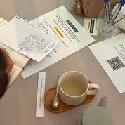 A top-down view of a desk with a white ceramic teapot, a white mug of tea on a wooden saucer, papers, sticky notes, pens, and a QR code, with part of a person's head and shoulder visible on the left.