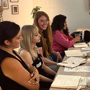 Four women seated at a long table in a meeting or workshop setting, with notebooks, papers, and a mug visible on the table. The room has warm lighting and framed pictures on the wall.