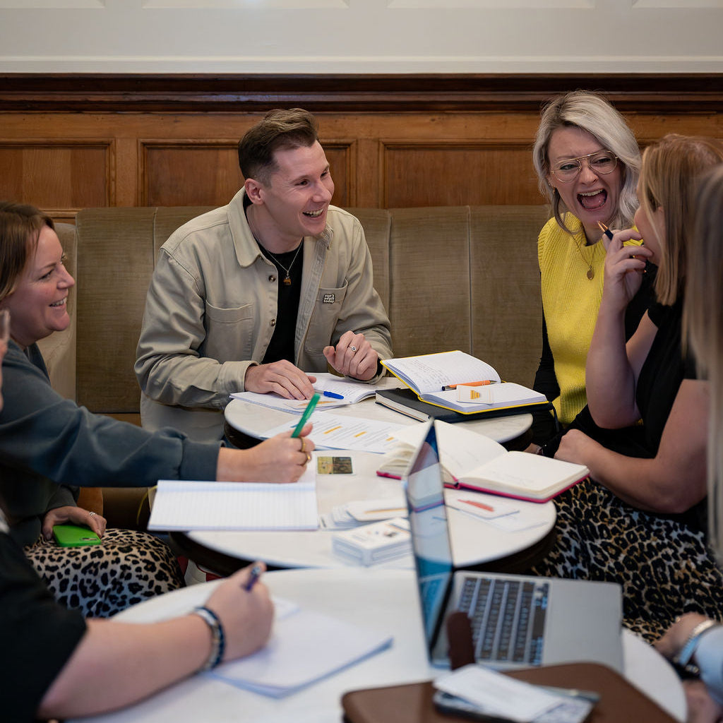 A group of four colleagues sitting around a round table in a warm, indoor office or cafe setting, laughing and writing in notebooks with laptops open on the table.
