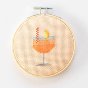 Cross-stitched design of a cocktail glass with a lemon wedge in a wooden hoop on a white background