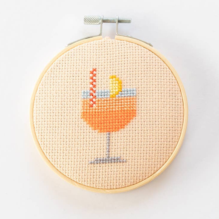 Cross-stitched design of a cocktail glass with a lemon wedge in a wooden hoop on a white background