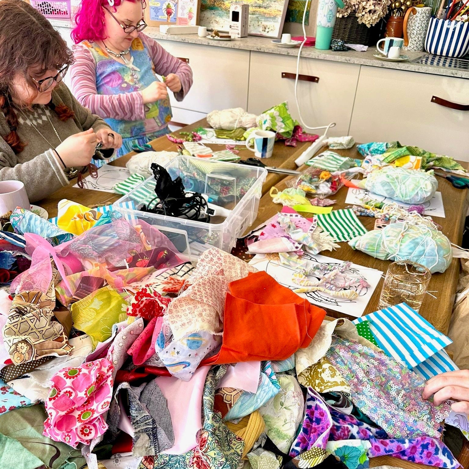 People working with fabric and sewing materials on a table in a kitchen.