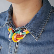 A close-up of a person wearing a chunky, multi-colored braided cord necklace with yellow, red, green, and white segments, styled over a blue denim shirt.