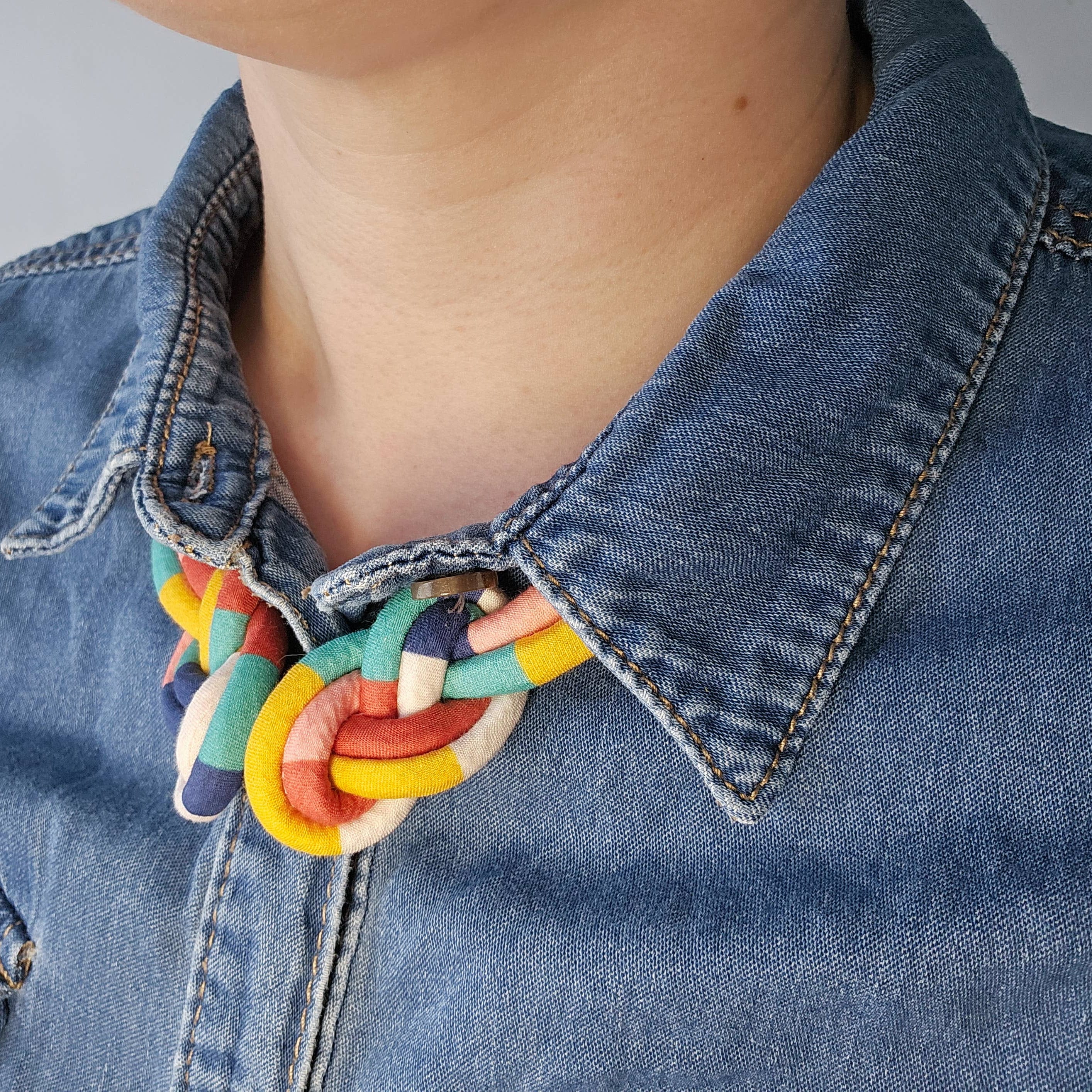 A close-up of a person wearing a chunky, multi-colored braided cord necklace with yellow, red, green, and white segments, styled over a blue denim shirt.