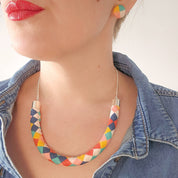 A close-up lifestyle shot of a woman wearing a colorful geometric chevron necklace and matching stud earrings, styled with a denim shirt against a plain light background.
