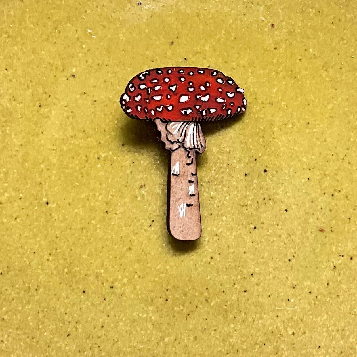 A small mushroom-shaped accessory, likely an enamel pin or charm, featuring a red cap with white spots and a pinkish-brown stem, centered on a round, speckled yellow-green ceramic plate against a neutral gray background.