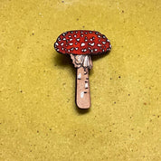 A small mushroom-shaped accessory, likely an enamel pin or charm, featuring a red cap with white spots and a pinkish-brown stem, centered on a round, speckled yellow-green ceramic plate against a neutral gray background.