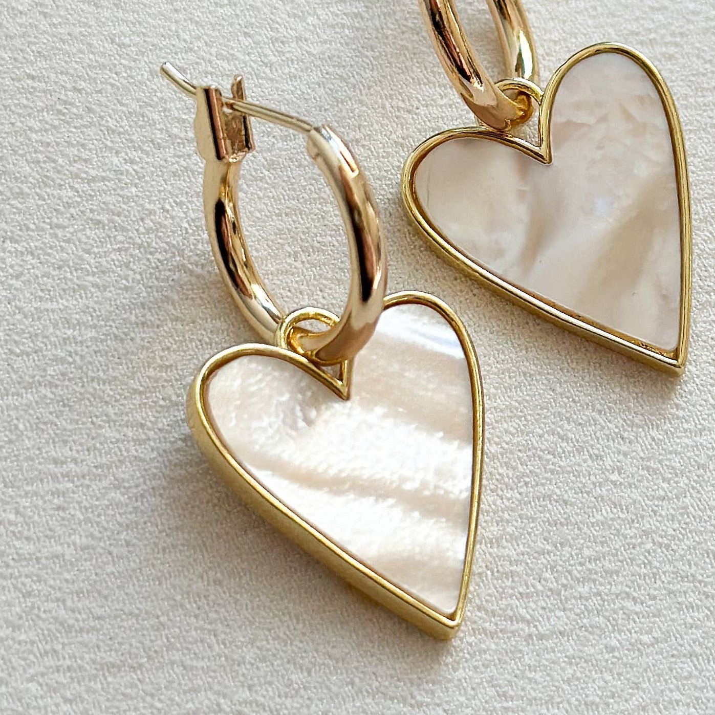 A pair of gold hoop earrings with heart-shaped pendants made of iridescent mother-of-pearl, displayed on a textured off-white surface.
