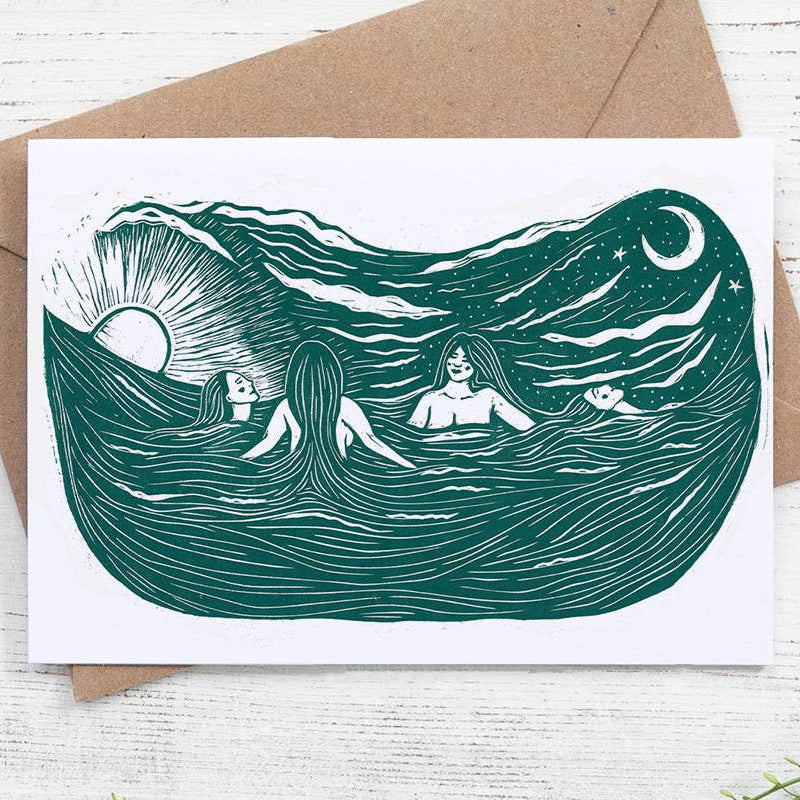 Greeting card with ocean design featuring people and a brown envelope on a white wooden surface.