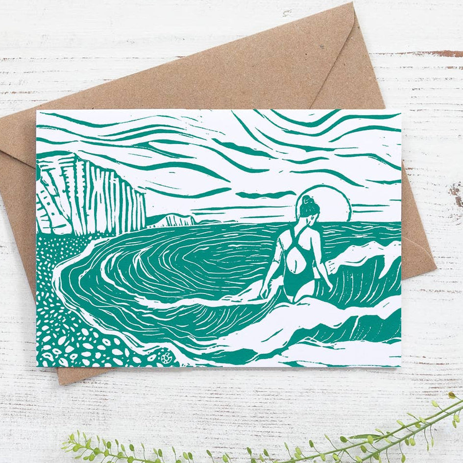 Greeting card with a design of a person in a swimsuit sitting on a wave, accompanied by a brown envelope on a white wooden surface.
