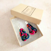 A pair of colorful fabric drop earrings with silver fishhook wires, presented in an open beige gift box with a branded lid on a light beige surface.