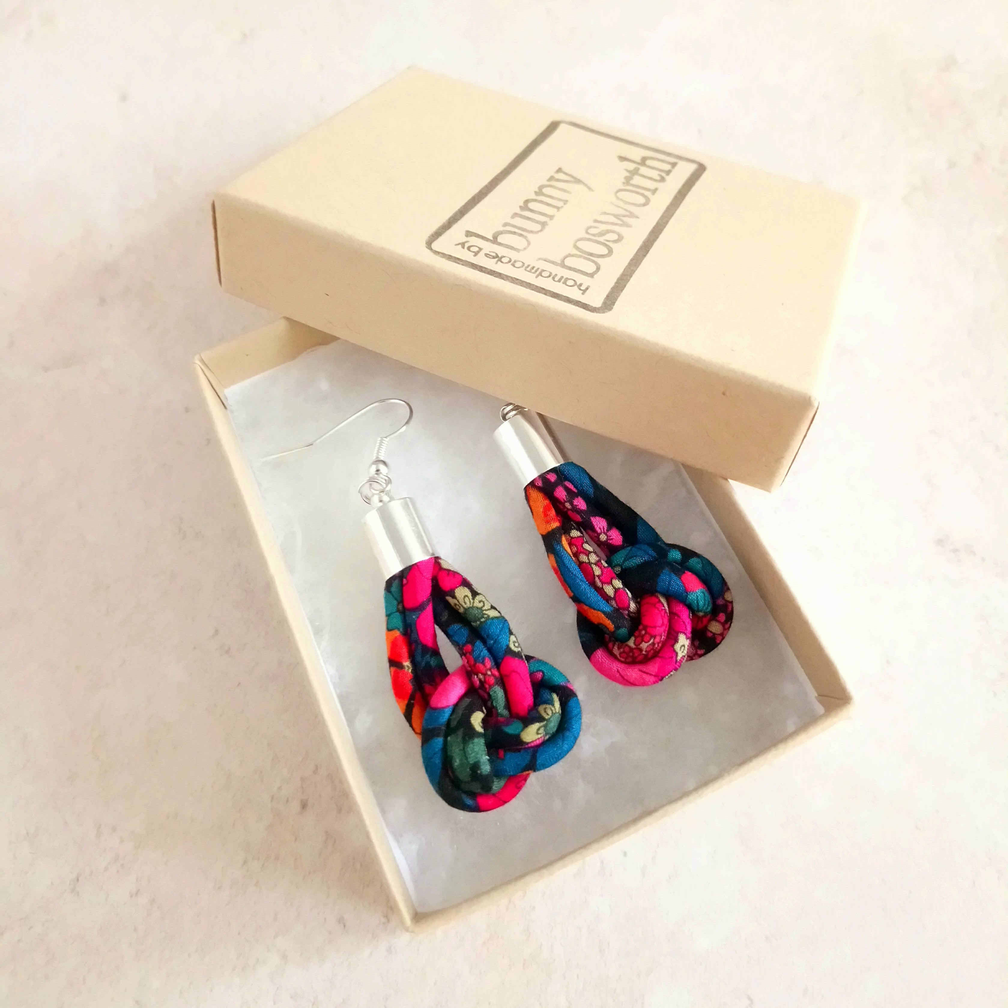 A pair of colorful fabric drop earrings with silver fishhook wires, presented in an open beige gift box with a branded lid on a light beige surface.