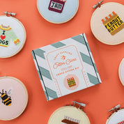 Collection of cross-stitched embroidery hoops on an orange background with a Cotton Clara box.