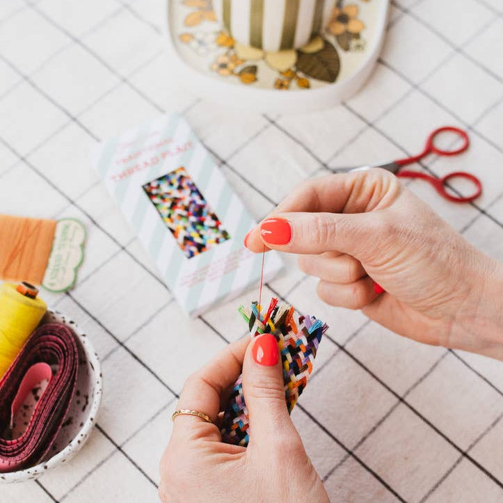 Person braiding colorful yarn on a tiled floor with a cup and scissors in the background.