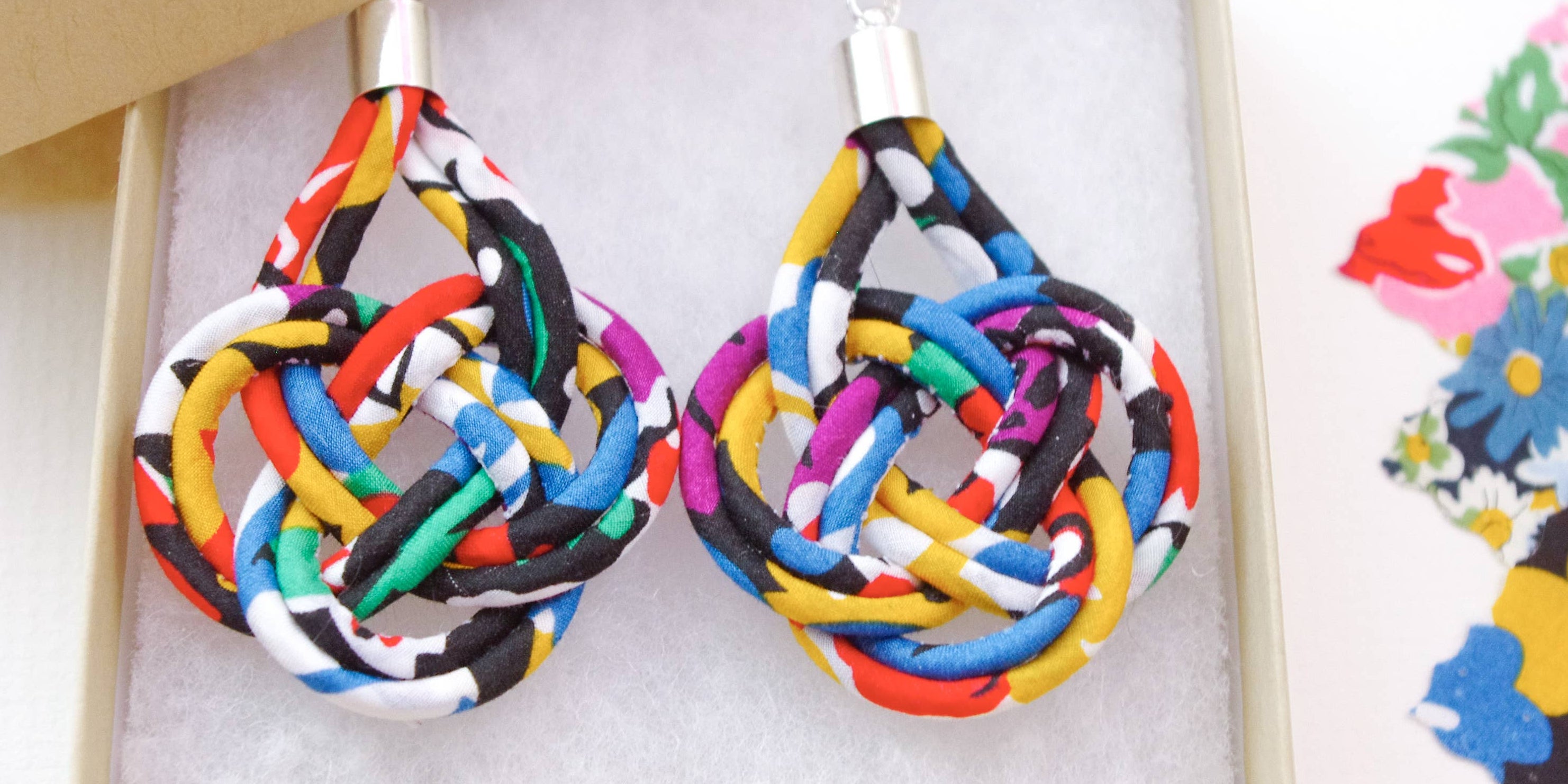 A pair of handmade teardrop-shaped earrings made from knotted multicolor fabric strips, presented in a light beige gift box with white foam insert. The box is shown on a white surface with sewing supplies, including floral fabric, scissors, and a spool of thread, in the background.