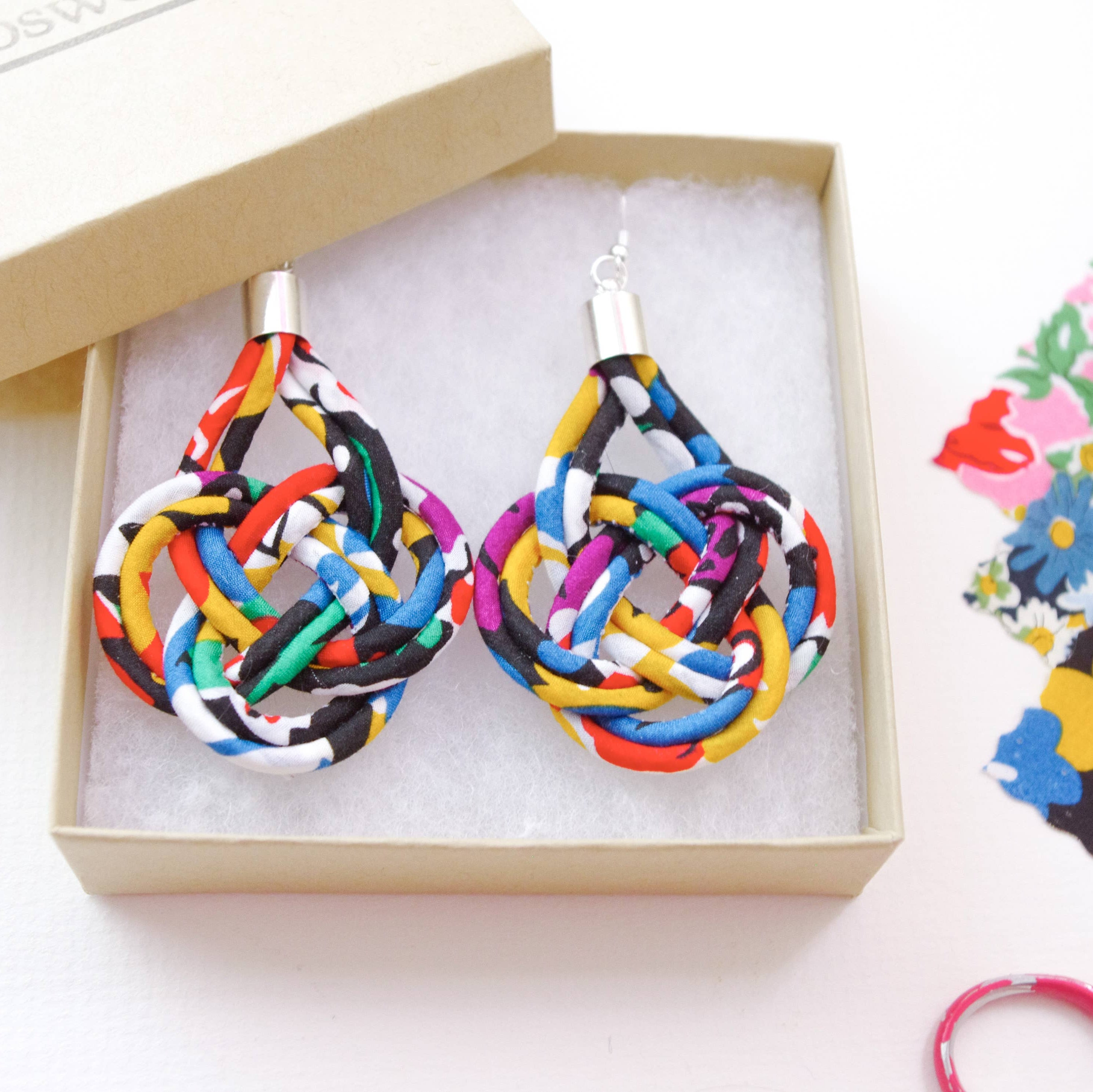 A pair of handmade teardrop-shaped earrings made from knotted multicolor fabric strips, presented in a light beige gift box with white foam insert. The box is shown on a white surface with sewing supplies, including floral fabric, scissors, and a spool of thread, in the background.