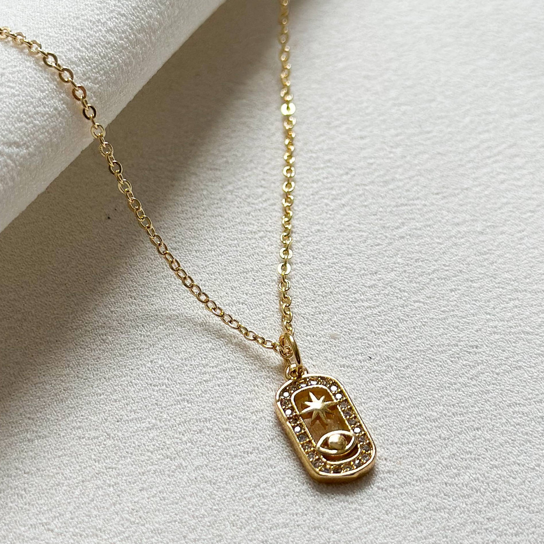 A delicate gold chain necklace with a small rectangular pendant featuring a stylized sun or moon face, displayed on a textured white fabric background.
