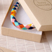 A braided multicolor fabric headband by bunny bosworth displayed inside its kraft paper gift box, with branded packaging cards and a stamp visible on a woven mat background.