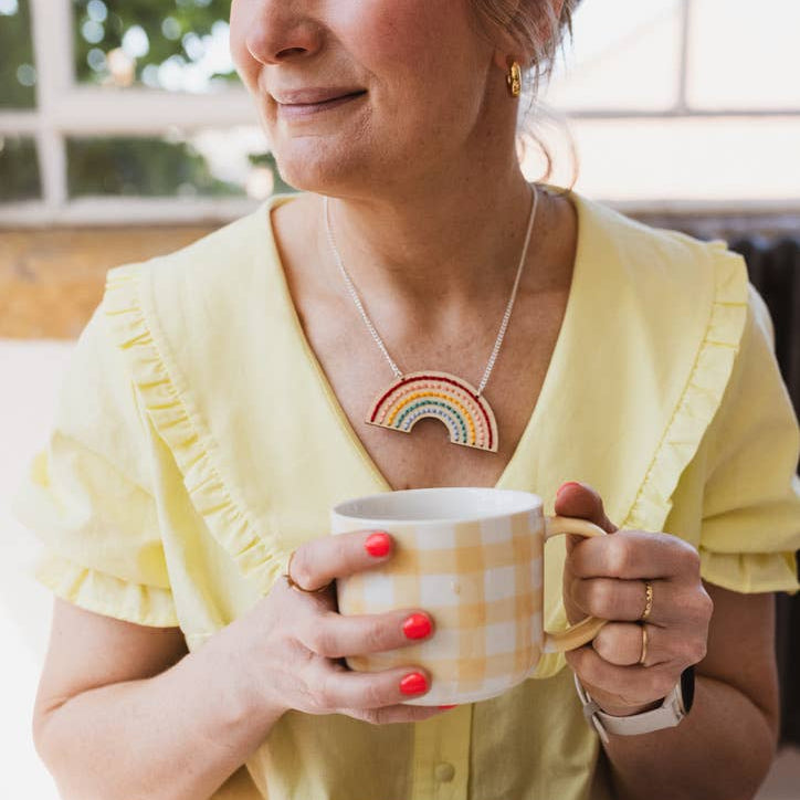 Woman in yellow shirt holding a mug with a rainbow necklace, sitting at a table.