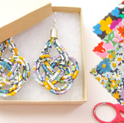 A pair of handmade teardrop-shaped dangle earrings made from knotted, multi-colored patterned ribbon, presented in a light wood box. The earrings feature a multicolor floral and geometric pattern with yellow, blue, purple, and white tones.