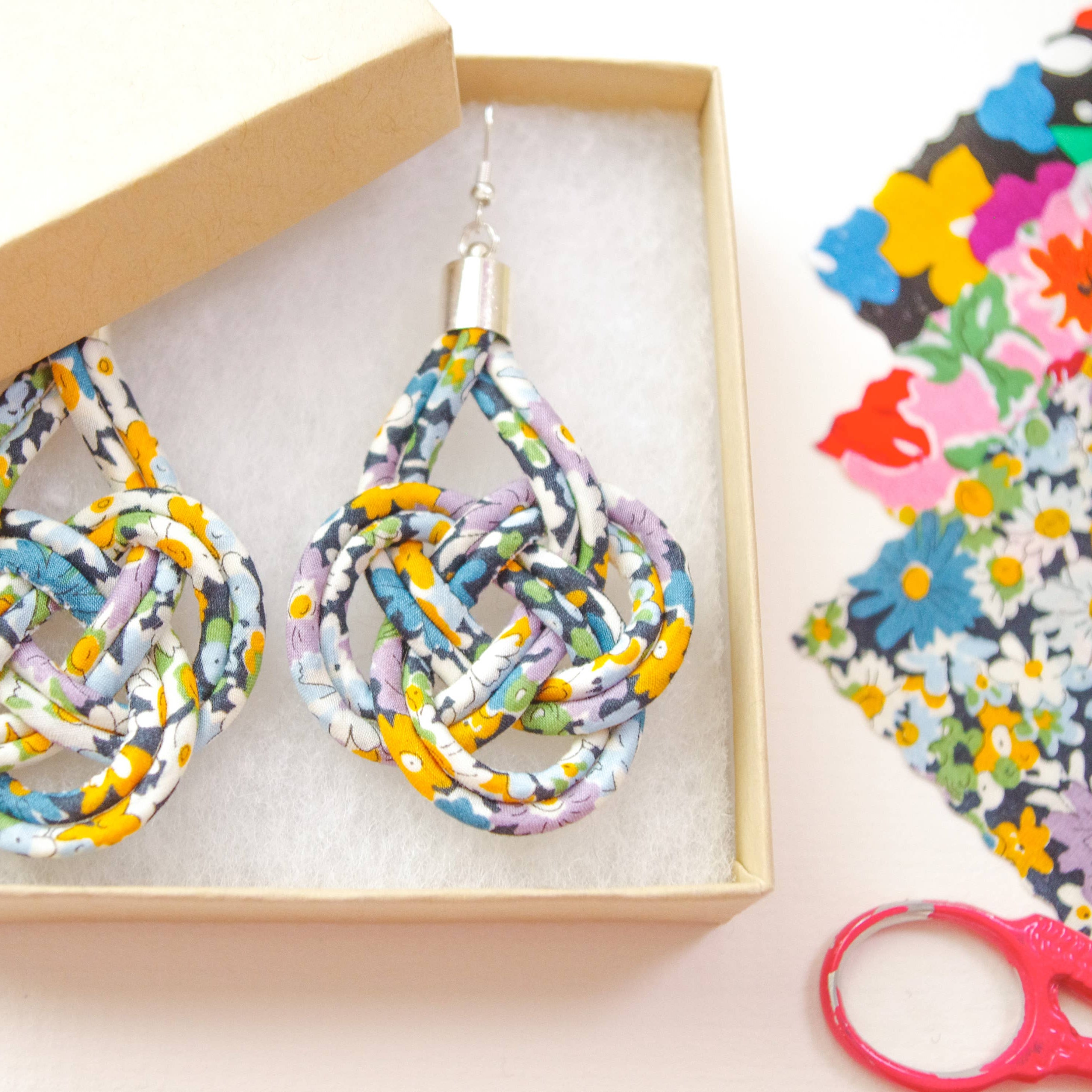 A pair of handmade teardrop-shaped dangle earrings made from knotted, multi-colored patterned ribbon, presented in a light wood box. The earrings feature a multicolor floral and geometric pattern with yellow, blue, purple, and white tones.