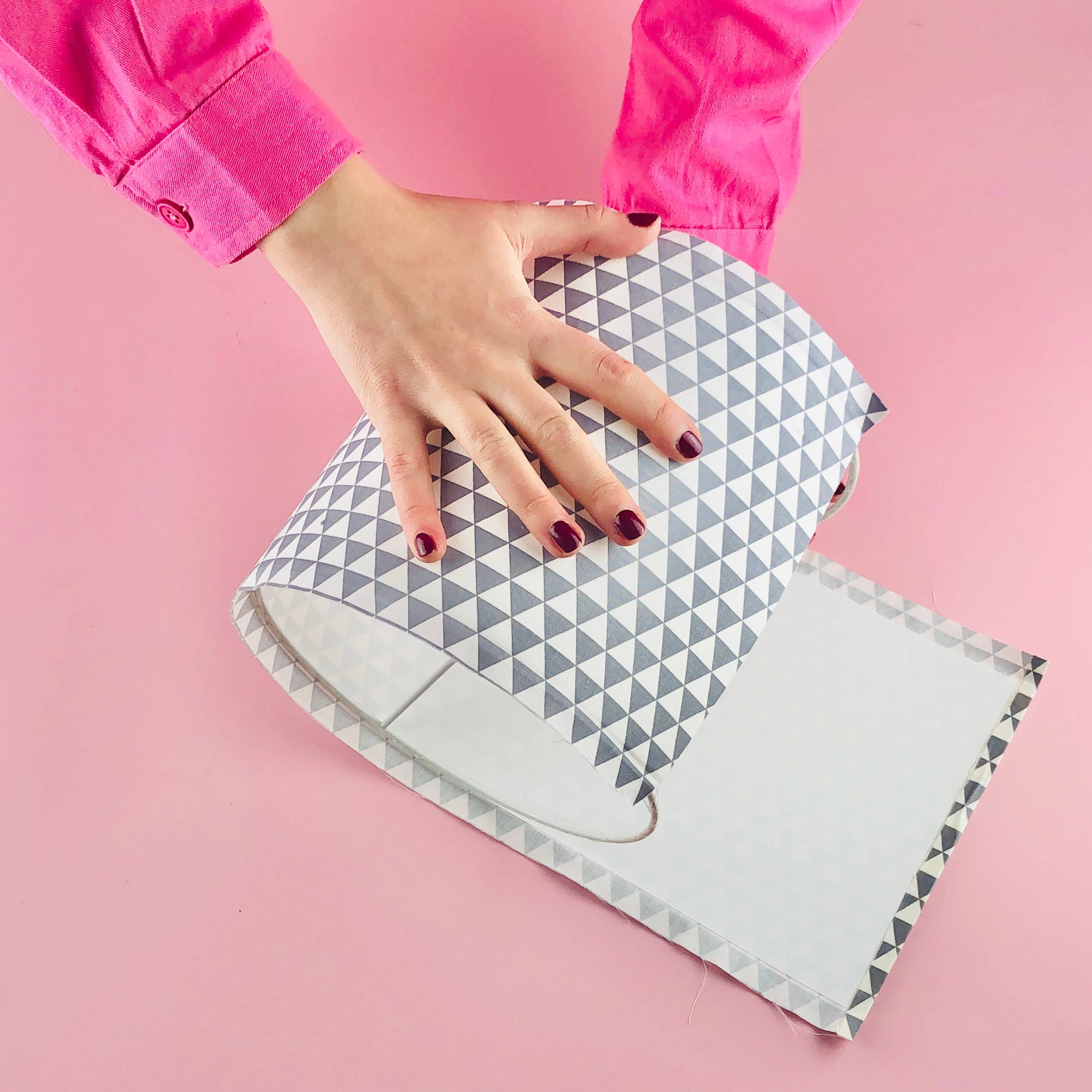 A person wearing a bright pink long-sleeve top is shown from the arms down, holding a rolled sheet of patterned vinyl or craft paper with a gray and white geometric triangle design on a solid pink background.