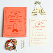 Cotton Clara embroidery kit with wooden necklace, yarn, and rainbow-shaped tool on a white background