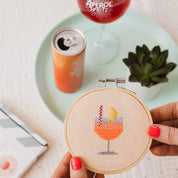 Hand holding a cross-stitched hoop with a cocktail design, surrounded by drinks and a plant on a table.