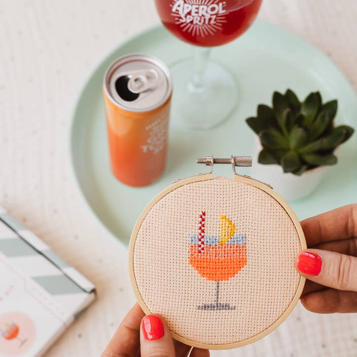 Hand holding a cross-stitched hoop with a cocktail design, surrounded by drinks and a plant on a table.