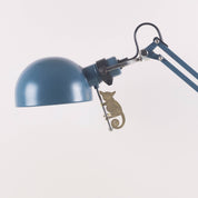 A blue adjustable desk lamp with a dome-shaped shade and articulated arm, featuring a small metallic cat-shaped accent near the joint, photographed against a plain white background.