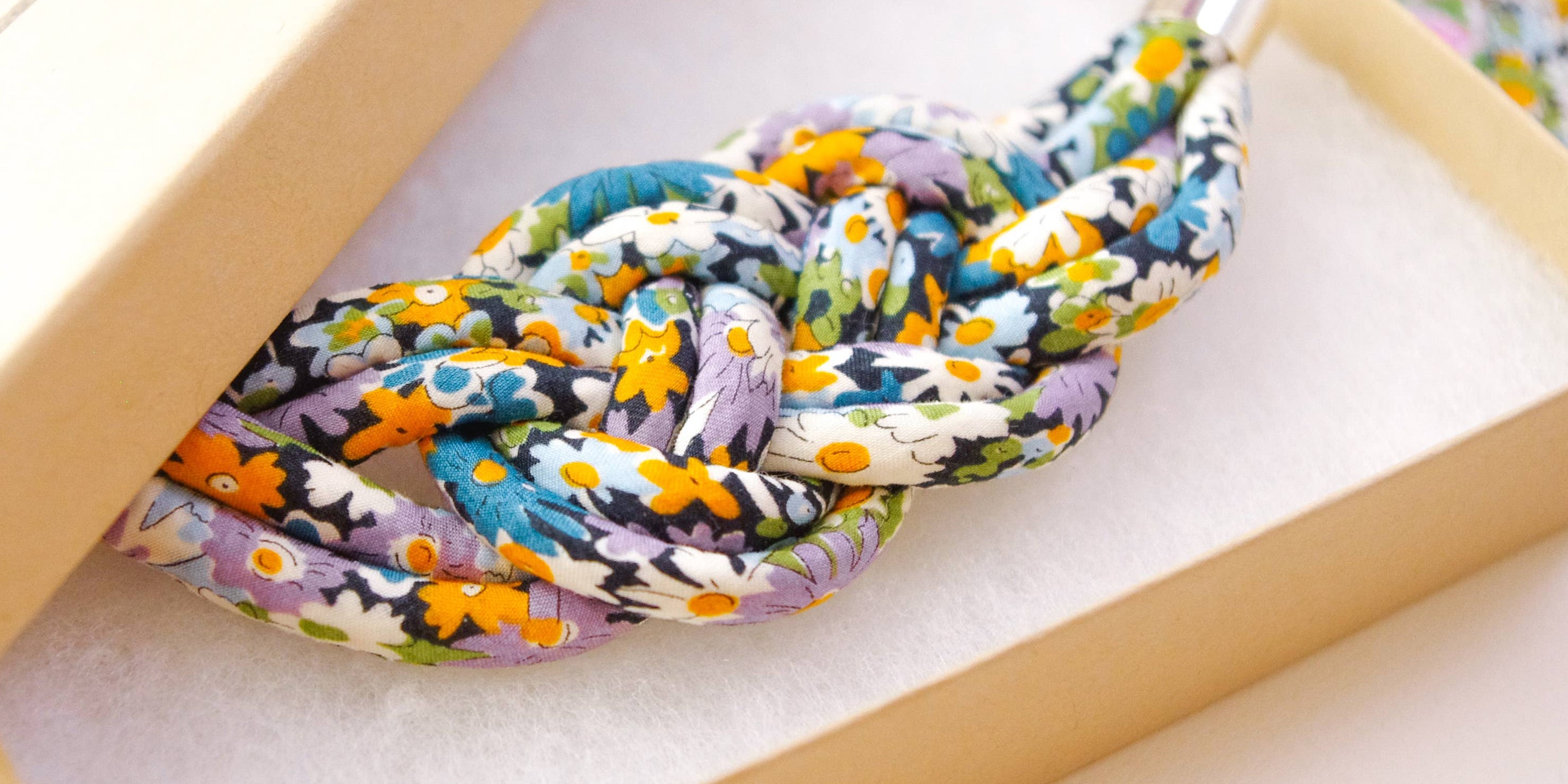 A knotted multicolor floral rope accessory displayed inside an open beige presentation box with a matching lid, photographed on a light neutral surface.