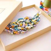 A knotted multicolor floral rope accessory displayed inside an open beige presentation box with a matching lid, photographed on a light neutral surface.