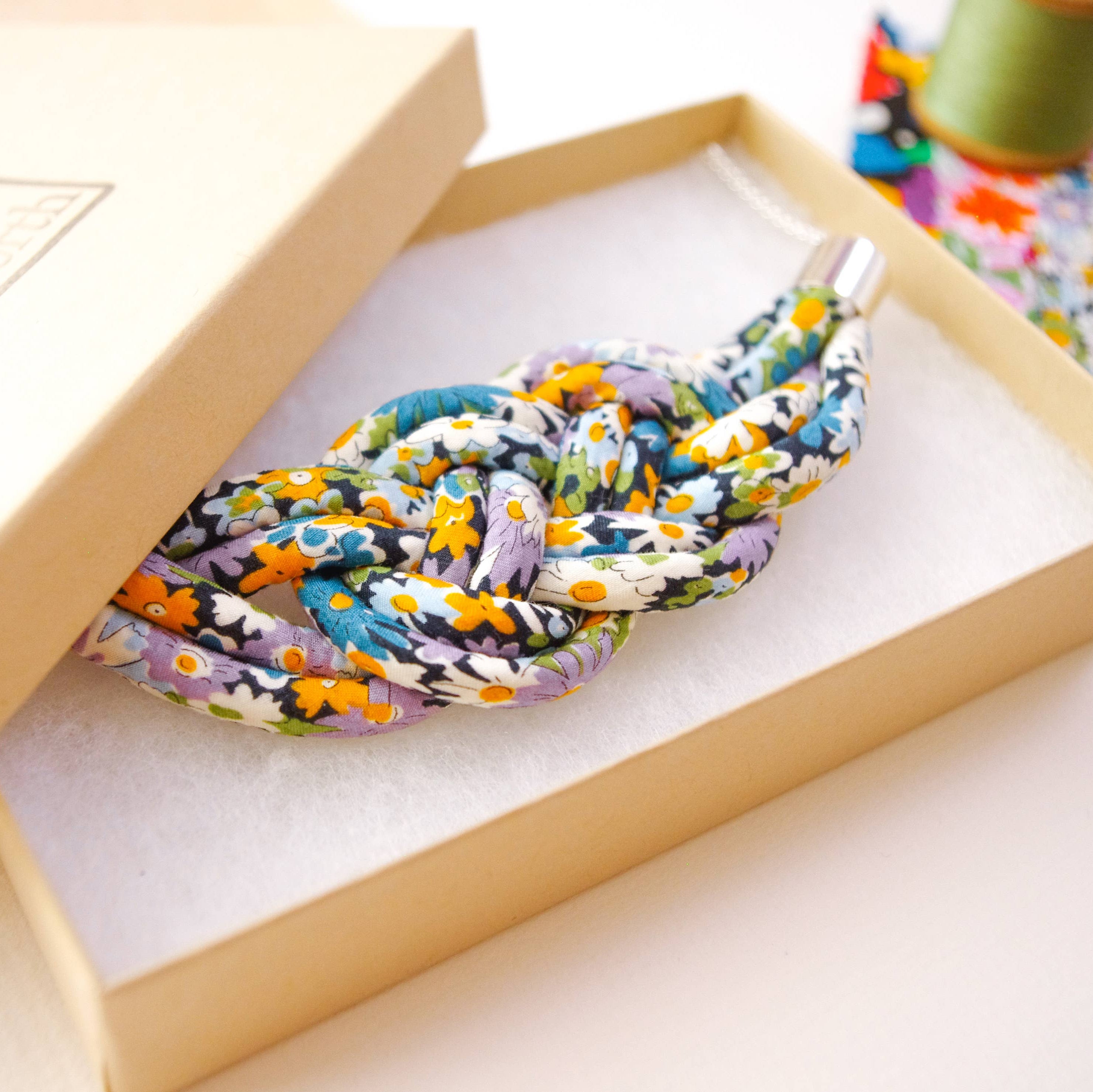A knotted multicolor floral rope accessory displayed inside an open beige presentation box with a matching lid, photographed on a light neutral surface.
