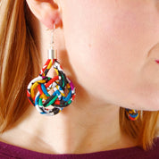 A close-up of a woman wearing colorful handmade earrings made from knotted ribbon strips, with her face partially visible in profile against a light background.