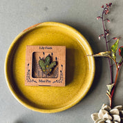 A small square wooden card labeled 'Lily Faith Mini Pin' sits in the center of a yellow ceramic plate on a gray surface, styled with a seashell and dried flowers.