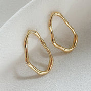 A pair of gold abstract open hoop earrings with a textured, organic wavy outline, displayed on a soft, draped white fabric background.