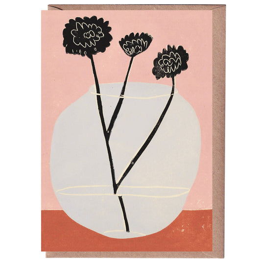 A greeting card with a linocut print of three black flowers in a white bowl, set against a pink background. The card has a kraft envelope.