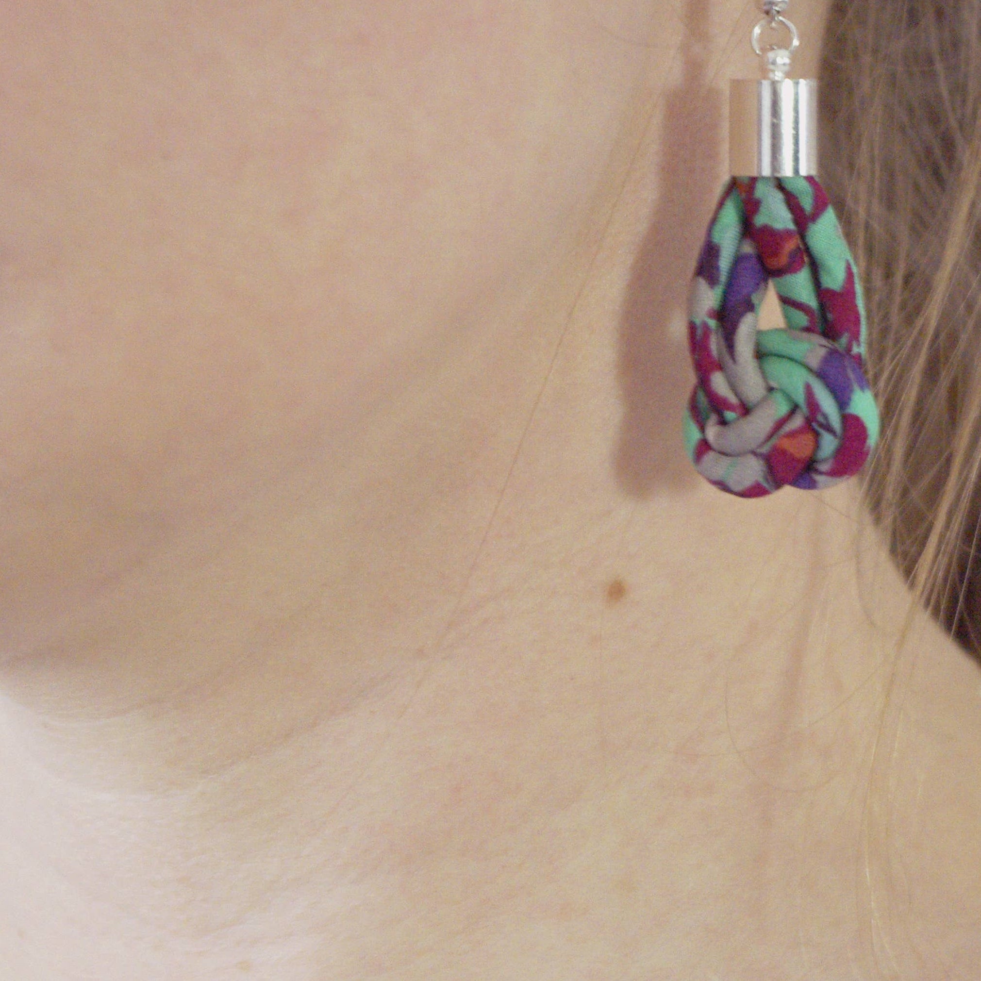 A close-up of a person's ear wearing a dangling earring made from colorful patterned fabric with a silver-toned metal top and hook.