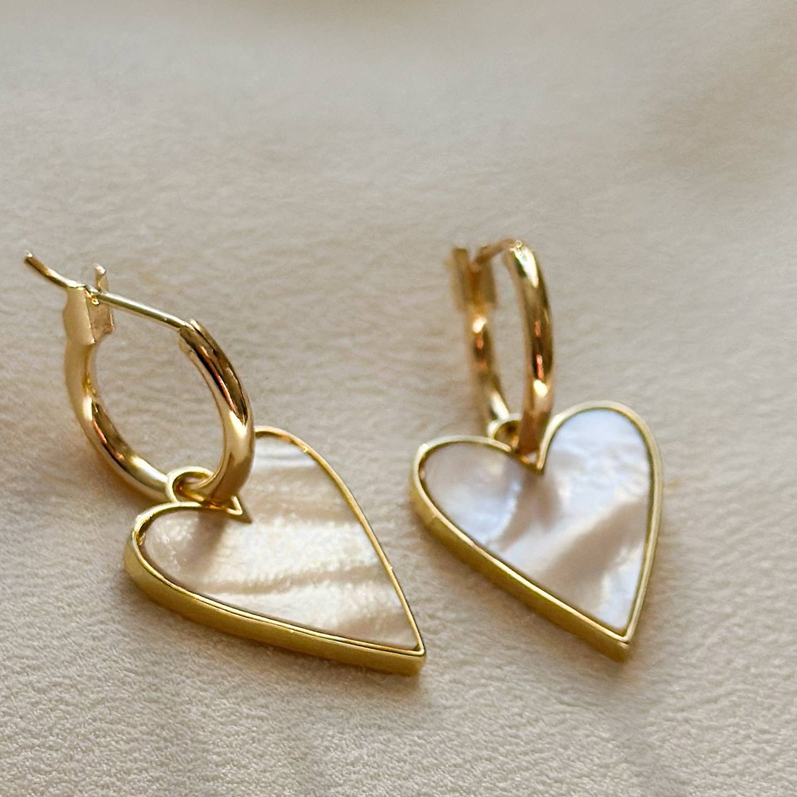 A pair of gold hoop earrings with dangling heart-shaped pendants featuring a pearlescent white inlay, displayed on a soft beige textured surface.
