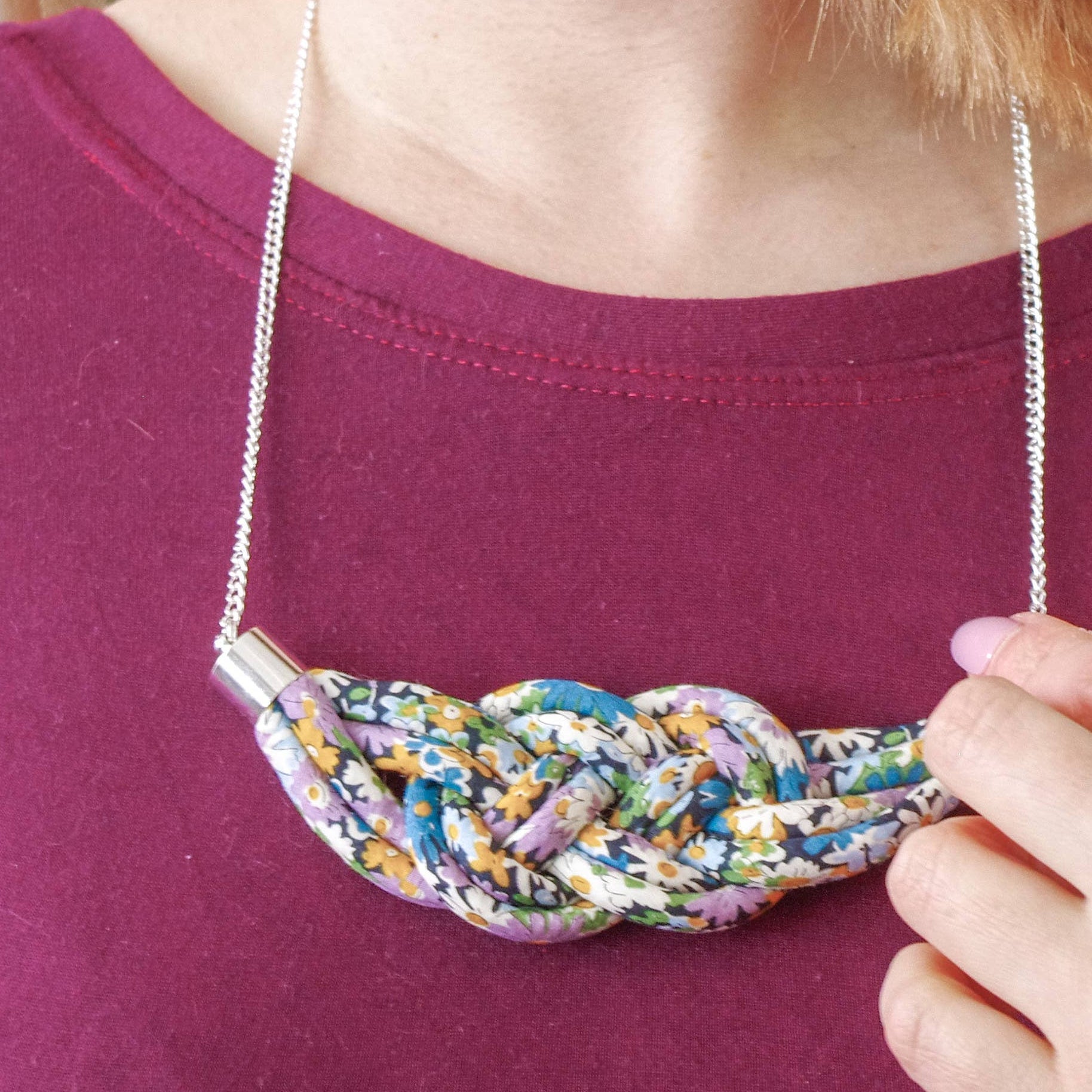 A close-up of a person wearing a chunky, braided statement necklace made from colorful shredded paper, styled with a silver chain over a magenta t-shirt.