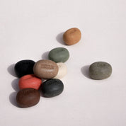 Nine smooth, oval-shaped stone pucks in various colors (black, brown, red, beige, green, gray, and white) arranged on a plain off-white background. Each stone features a debossed brand name starting with 'PUL'.