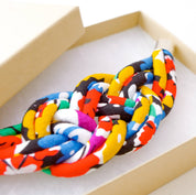 A colorful knotted fabric accessory with a bold abstract pattern in red, yellow, blue, green, black, and white, presented inside a light beige cardboard box.