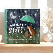 A children's book titled "Watching from the Stars" by Shrey McKee sits on a light wood shelf in a softly lit room. The cover shows a dark rainy night sky with stars and a crescent moon, a fox holding a green umbrella, and a small dog on a blanket.