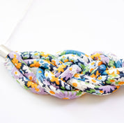 A close-up of a braided fabric necklace with a colorful floral pattern in purple, orange, blue, green, and white, threaded through silver metal spacers on a white background.