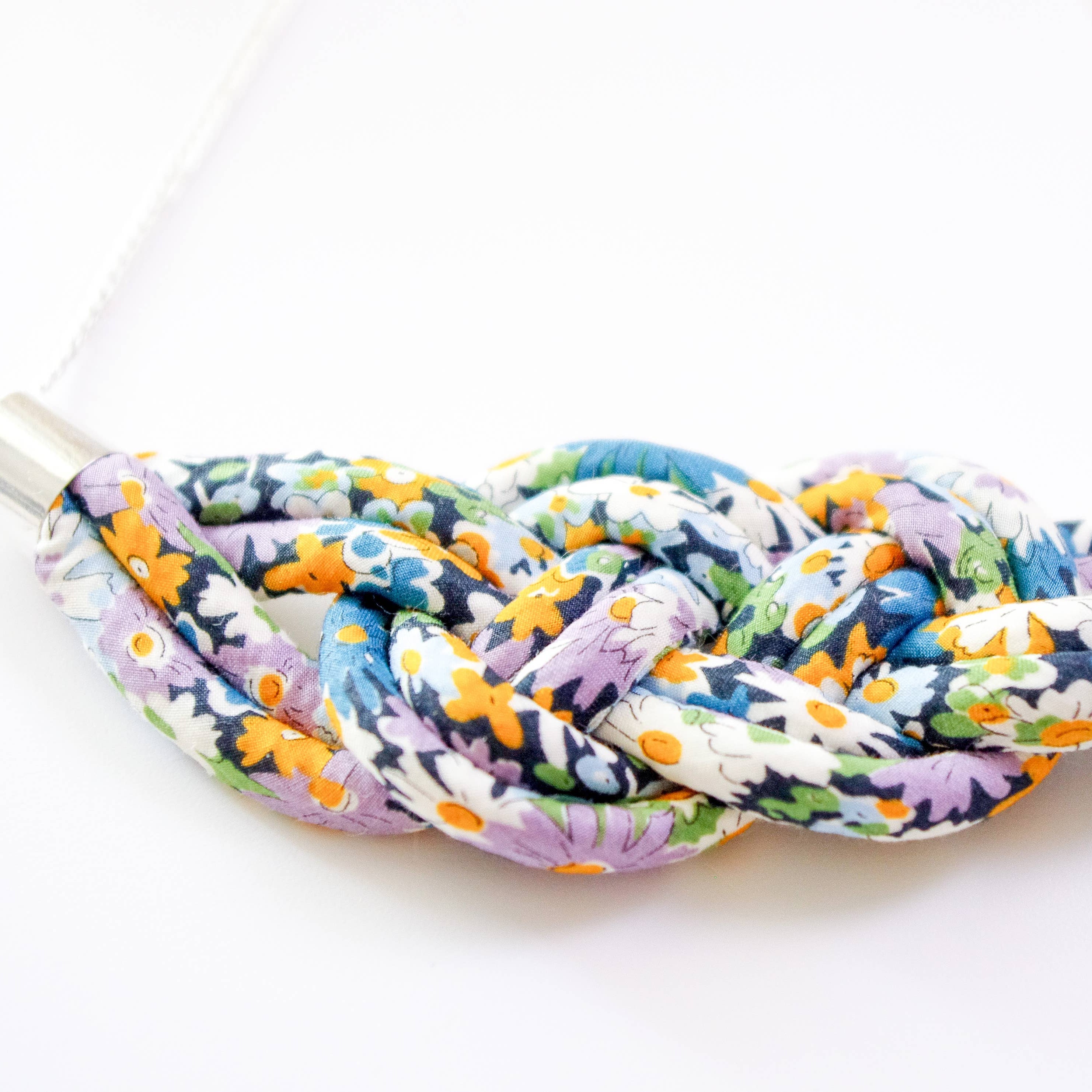 A close-up of a braided fabric necklace with a colorful floral pattern in purple, orange, blue, green, and white, threaded through silver metal spacers on a white background.