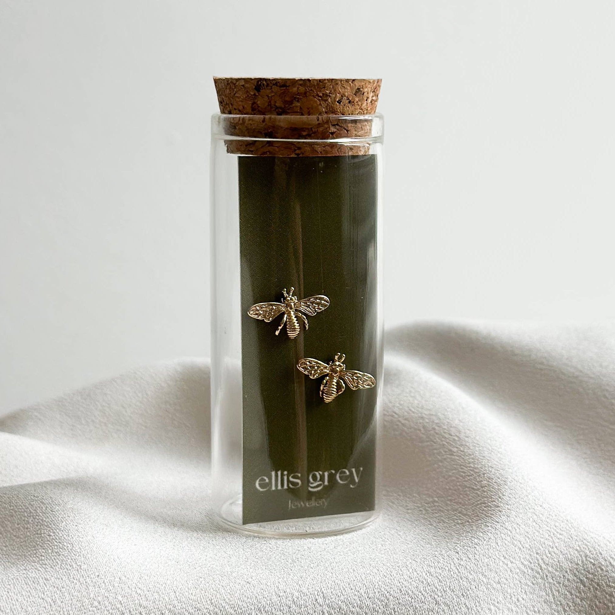 A pair of gold bee stud earrings displayed inside a clear glass vial with a cork stopper and a dark olive-green insert card. The card features the brand name 'ellis grey' in white text and is set against a draped white fabric background.